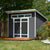 Modern backyard shed with a flat roof, transom windows, and double doors, surrounded by plants and a wooden deck.