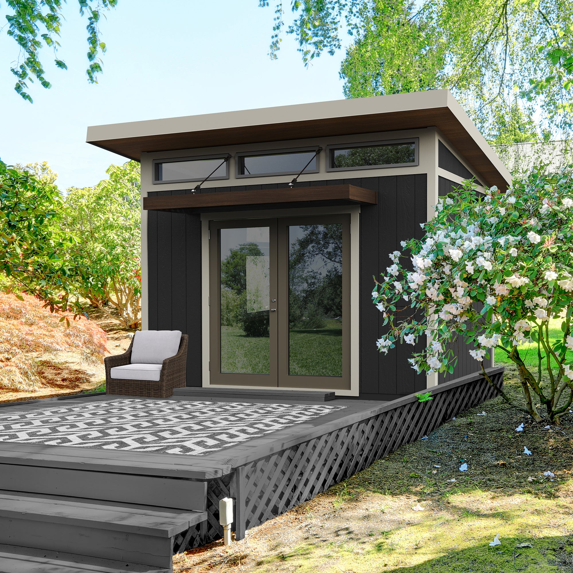 Modern studio shed with glass double doors, transom windows, and a wooden overhang on a raised deck with seating.