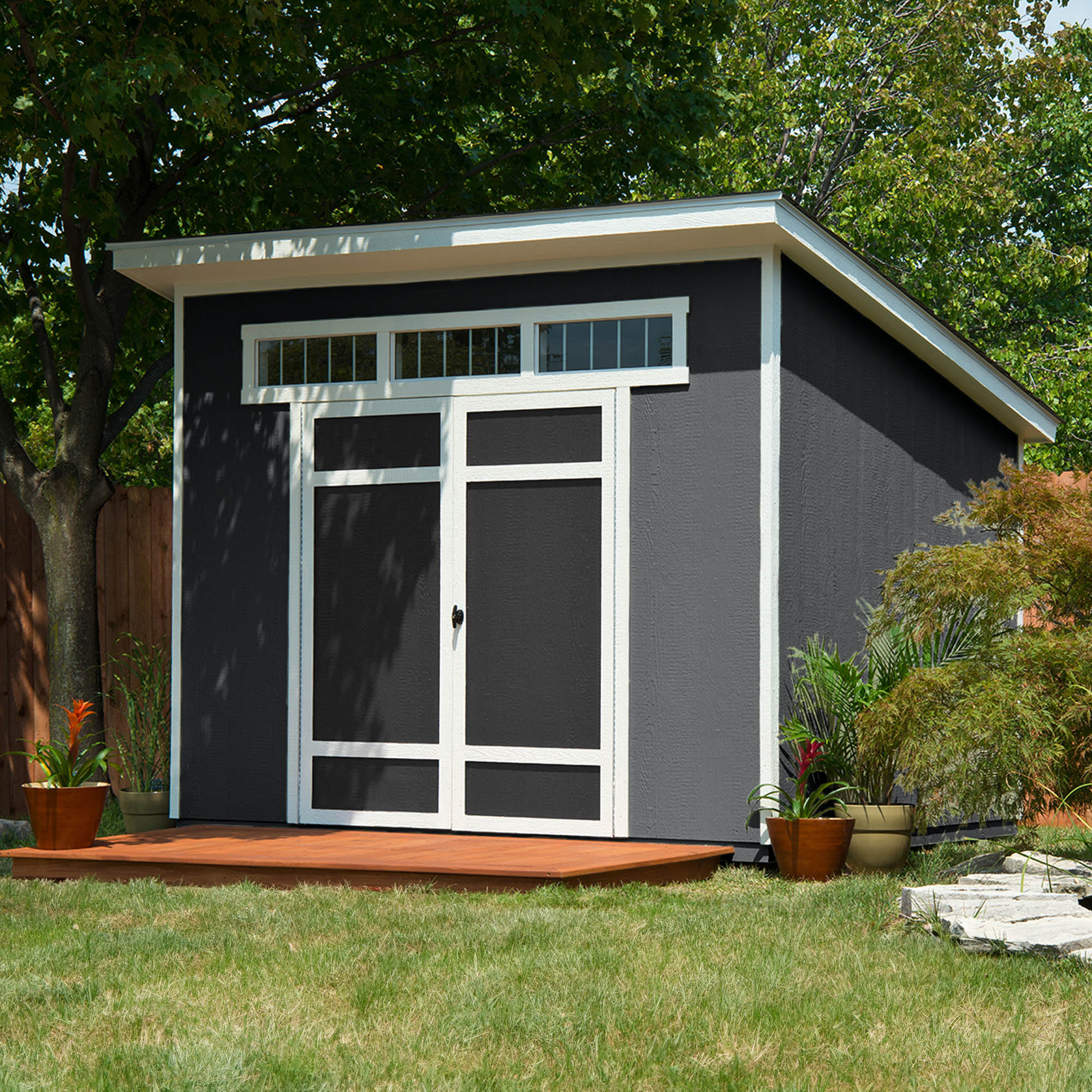 Modern backyard shed with a flat roof, transom windows, and double doors, surrounded by plants and a wooden deck.